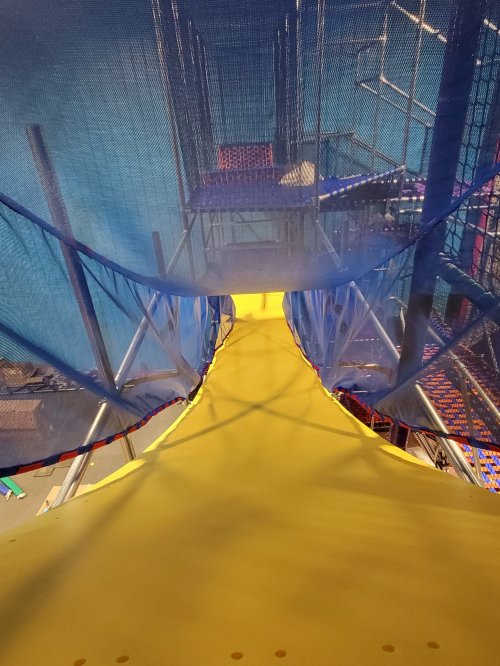 View from the top of a yellow slide, leading down into a colorful play area surrounded by safety nets.