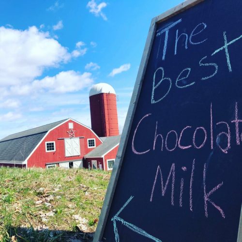 A red barn and silo under a blue sky, with a chalkboard sign pointing to "The Best Chocolate Milk."