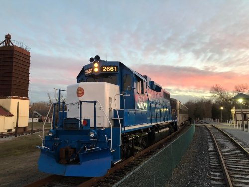 A blue and white train engine numbered 2661 parked on tracks at sunset, with a colorful sky in the background.