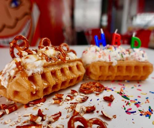 Two decorated waffles with ice cream, caramel, pretzels, and colorful sprinkles, featuring birthday candles spelling "HBD."