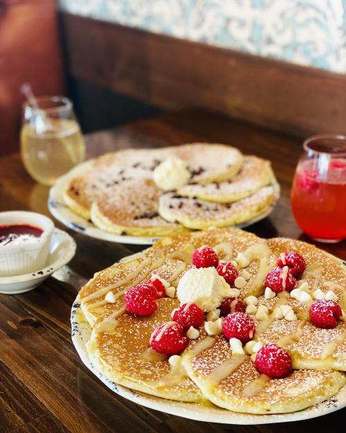 Two plates of pancakes topped with raspberries, whipped cream, and drizzled with syrup, served with drinks.