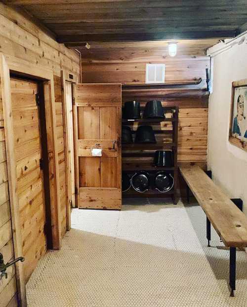 A wooden hallway with doors, a bench, and shelves holding black buckets in a rustic setting.