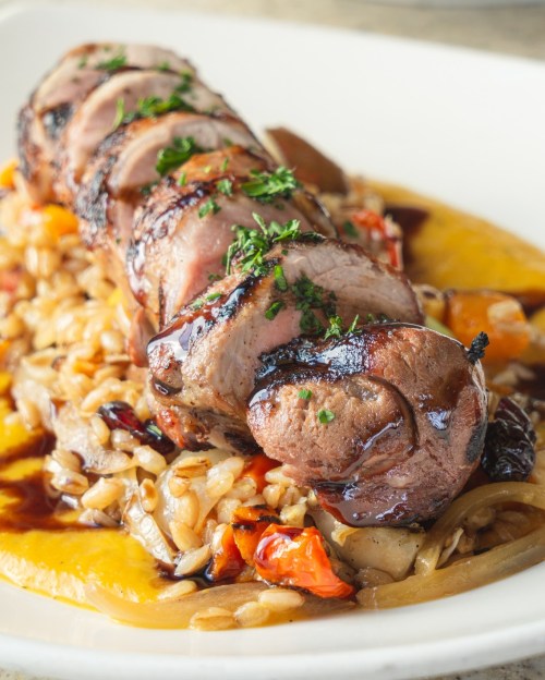 Sliced roasted meat on a bed of grains and vegetables, garnished with herbs and drizzled with sauce.