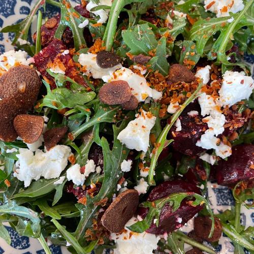 A vibrant salad with arugula, beets, crumbled feta, sliced almonds, and a sprinkle of chili flakes on a decorative plate.