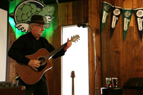 A musician in a hat plays an acoustic guitar on stage, with a microphone and drinks in the background.