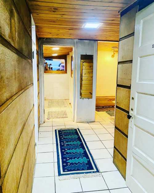 A hallway with wooden walls, tiled floor, and a patterned rug, leading to a well-lit room at the end.