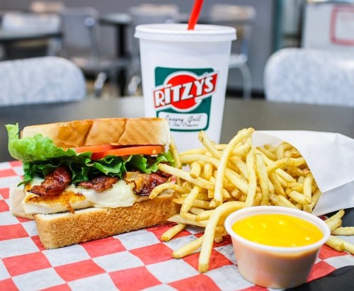 A sandwich with lettuce, tomato, and bacon, served with fries and a cup of yellow dipping sauce, alongside a drink.