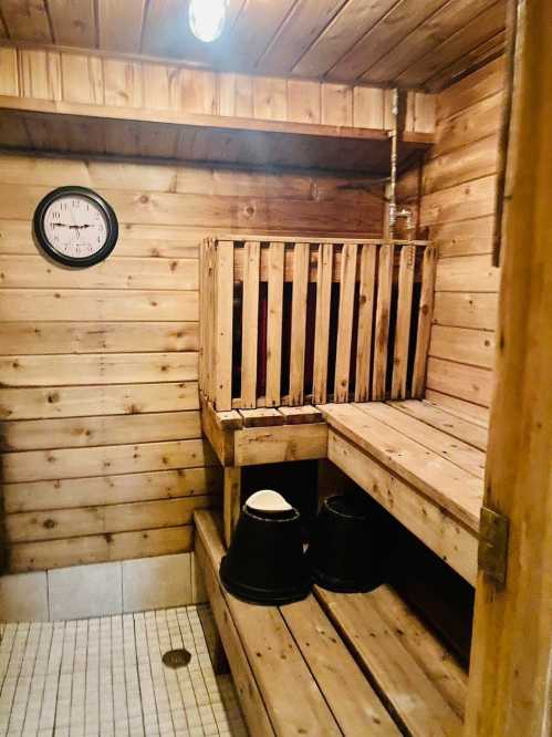 A wooden sauna interior featuring benches, a clock, and two black buckets on the floor.