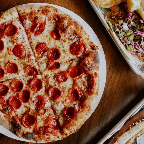 A round pepperoni pizza with slices cut, served on a white plate, alongside a colorful salad and other dishes.