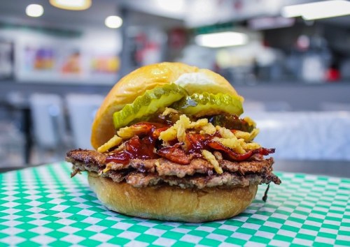 A close-up of a burger with a beef patty, bacon, pickles, and barbecue sauce on a toasted bun, set on a green checkered table.