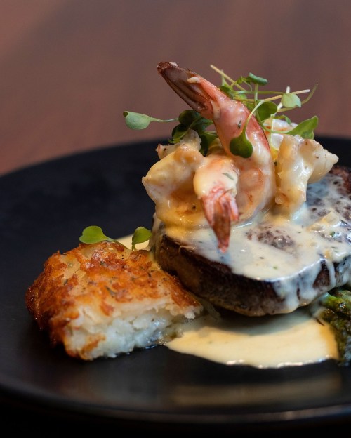 A gourmet dish featuring shrimp atop a steak with creamy sauce, served with a crispy potato cake and garnished with microgreens.