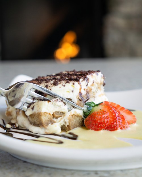 A slice of tiramisu topped with cocoa powder, served with a strawberry and a drizzle of cream on a white plate.