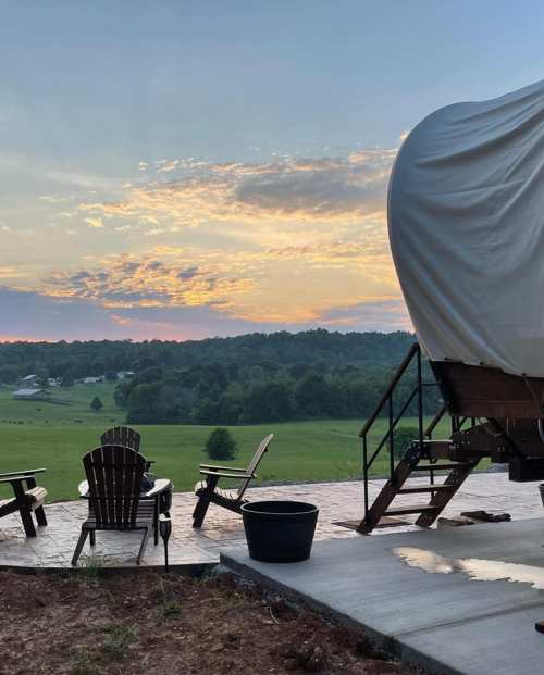 A serene outdoor scene with chairs facing a sunset over rolling hills and a covered structure nearby.