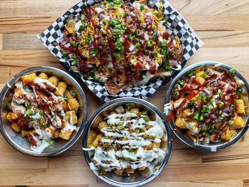 A variety of loaded tater tots in metal bowls, topped with cheese, sauces, and fresh vegetables on a wooden table.