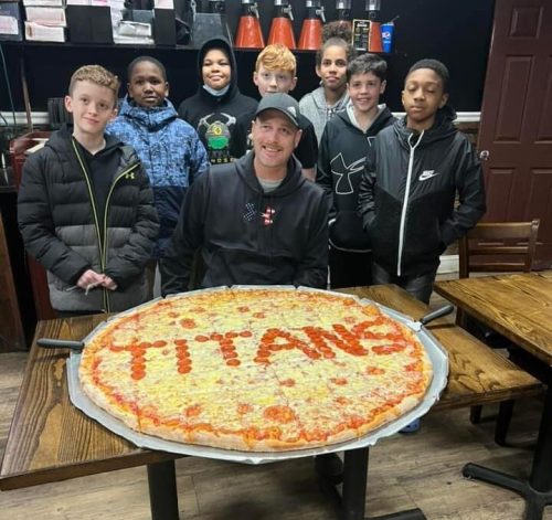 A group of kids poses with a large pizza labeled "TITANS" in a restaurant.