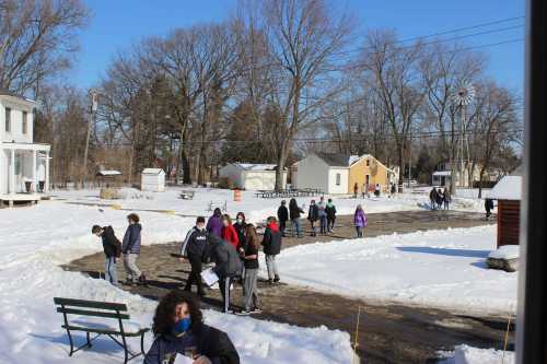 A snowy outdoor scene with children playing and walking on a path, surrounded by trees and buildings.