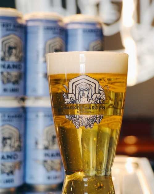 A glass of golden beer with a frothy head, featuring a logo, in front of stacked cans.