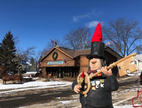 A cheerful statue in a red hat holding a guitar stands in front of a wooden building decorated for the holidays.