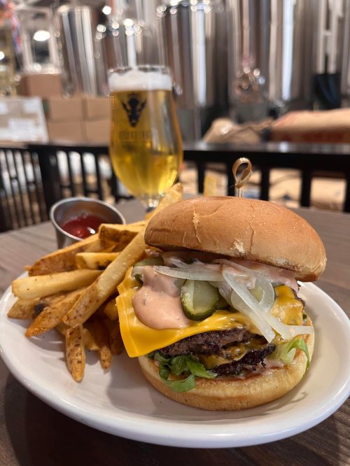 A juicy cheeseburger with lettuce, pickles, and sauce, served with fries and a glass of beer in a brewery setting.
