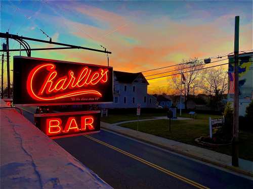 Neon sign for Charlie's Bar at sunset, with a colorful sky and a quiet street in the background.