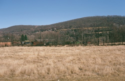 A wide, grassy field in the foreground with a distant, wooded hill under a clear blue sky.