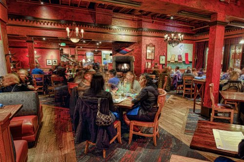 A bustling restaurant interior with diners at tables, warm lighting, and a cozy fireplace in the background.