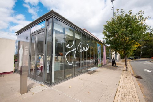 Modern glass building with signage, surrounded by trees and a sidewalk, on a cloudy day.