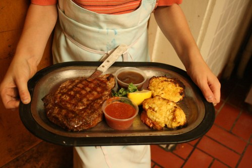 A person holds a metal tray with a grilled steak, two sides, and dipping sauces, set against a rustic background.