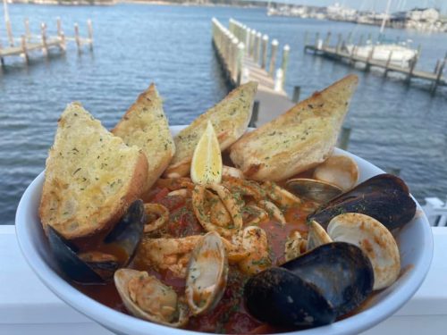 A bowl of seafood stew with garlic bread, set against a waterfront view with boats in the background.