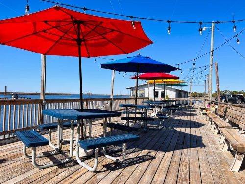 A wooden deck by the water with colorful umbrellas over picnic tables and string lights above. Clear blue sky.