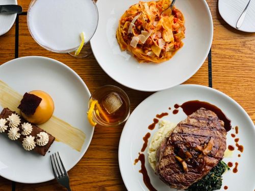 A table spread featuring steak with mashed potatoes, pasta, dessert, and a cocktail.