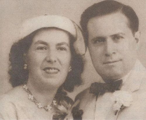 A vintage black-and-white photo of a smiling couple, dressed elegantly, with the woman wearing a hat and floral accessories.