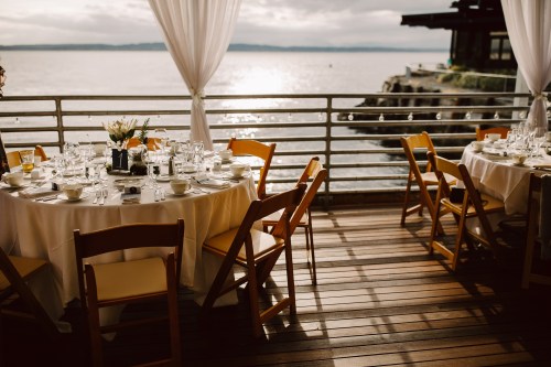 A beautifully set dining area with tables and chairs overlooking a serene waterfront at sunset.