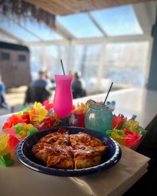 A plate of pizza slices with colorful tropical drinks and a floral lei in a bright, sunny setting.