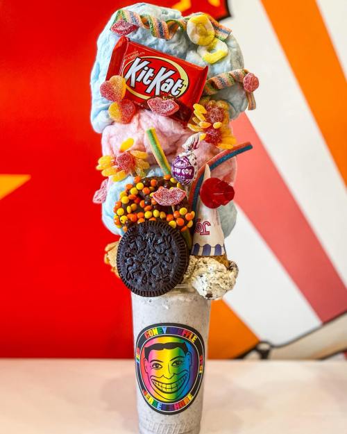 A colorful dessert topped with cotton candy, candy, a Kit Kat, and an Oreo in a cup with a smiling face design.