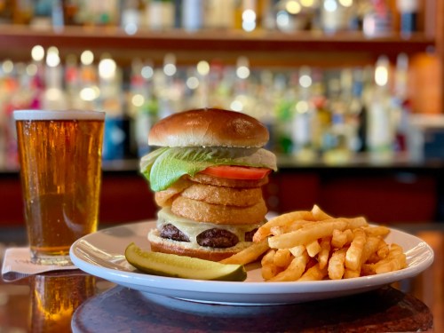 A towering burger with lettuce, tomato, and onion rings, served with fries and a glass of beer on a bar counter.