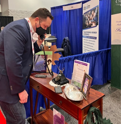 A man in a suit examines an antique phone on a table at a trade show booth, wearing a mask.