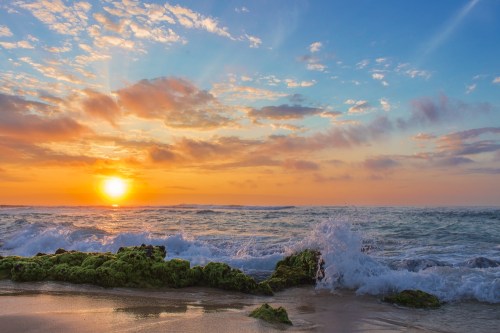 A vibrant sunset over the ocean, with waves crashing against mossy rocks and colorful clouds in the sky.