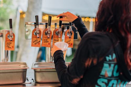A person fills a cup from a tap with various labeled drink options at a festival or outdoor event.