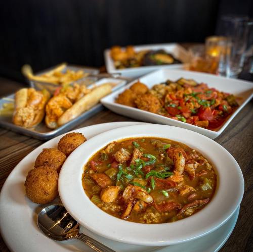 A bowl of shrimp gumbo garnished with green onions, surrounded by various side dishes on a wooden table.
