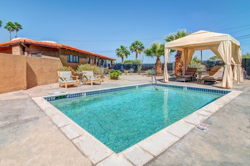 A clear swimming pool surrounded by palm trees and lounge chairs, with a shaded cabana in a sunny outdoor setting.
