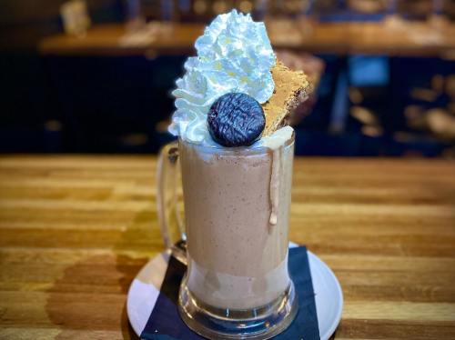 A tall glass of milkshake topped with whipped cream, a cookie, and a chocolate wafer on a wooden table.