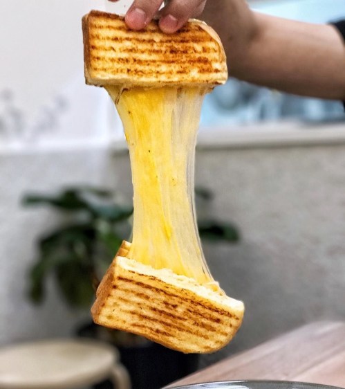 A hand holds a grilled cheese sandwich, showcasing gooey, melted cheese stretching between two toasted slices.