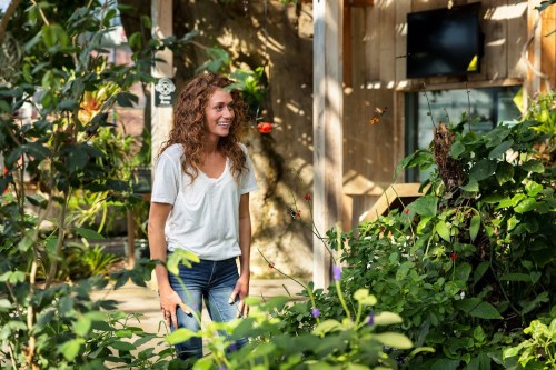 A smiling woman with curly hair stands among lush greenery in a bright, sunlit garden.