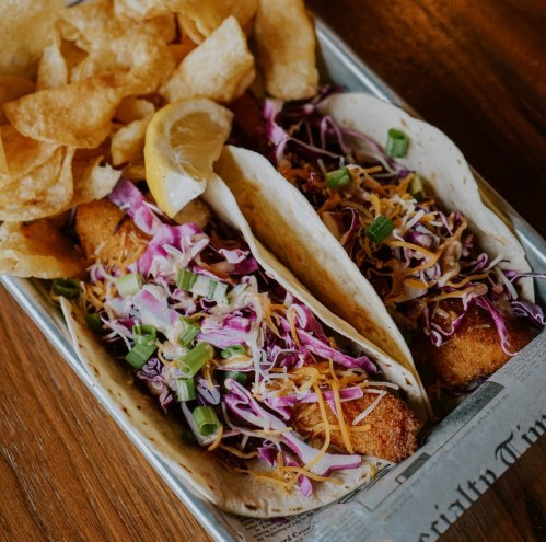 Two tacos filled with slaw, cheese, and crispy filling, served with potato chips and lemon wedges on a tray.