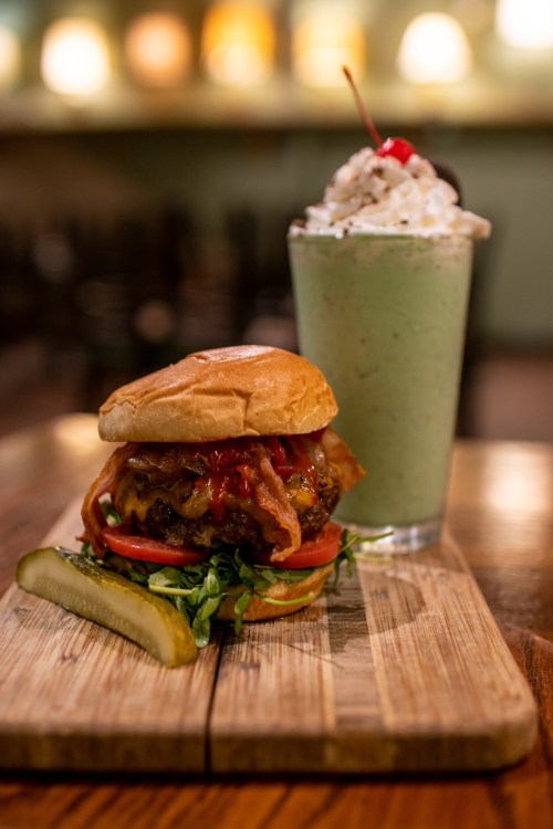 A juicy burger with lettuce, tomato, and bacon, served with a green milkshake topped with whipped cream and a cherry.
