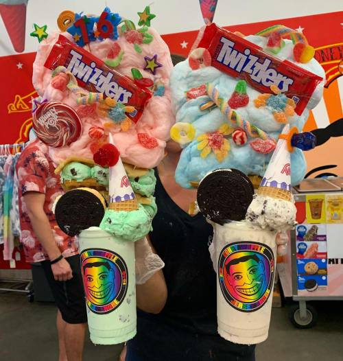 A person holds two oversized milkshakes topped with cotton candy, candy, and Twizzlers in a colorful ice cream shop.