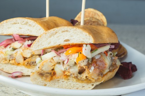 A close-up of a sandwich filled with grilled meat, pickled vegetables, and served on a toasted bun with chips on the side.