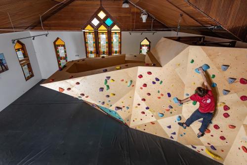 A person climbs a colorful bouldering wall inside a spacious, well-lit gym with stained glass windows.