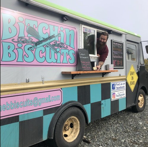 A man serves food from a colorful food truck named "Bitchin' Biscuits," with a menu displayed on the side.